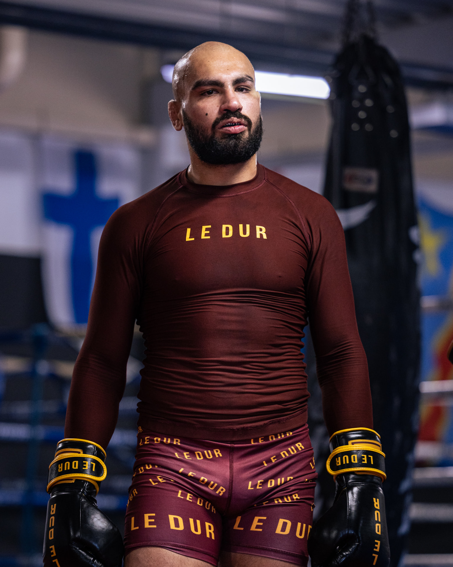 Lemar Khan standing in a gym, wearing a maroon LE DUR rash guard, maroon LE DUR shorts and black LE DUR boxing gloves with yellow straps.